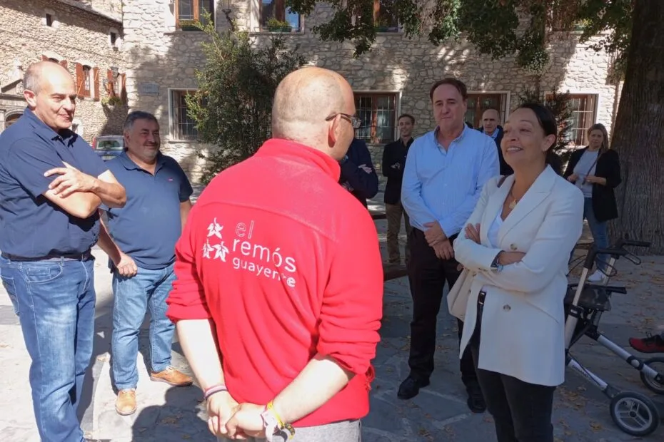 La consejera Carmen Susín charla con Manuel, que está de espalada y viste la sudadera roja de el Remos. Junto a la consejera se ve, de izquierda a derecha, a Aurelio Garcia, de la junta directiva de la asociacion guayente, Gaspar Mora, alcalde de Sesue, y Hector Rodriguez, director de el centro el remos. Detrás se ven oitras personas. Están junto al gran abeto de la plaza mayor de la Villa de Benasque.