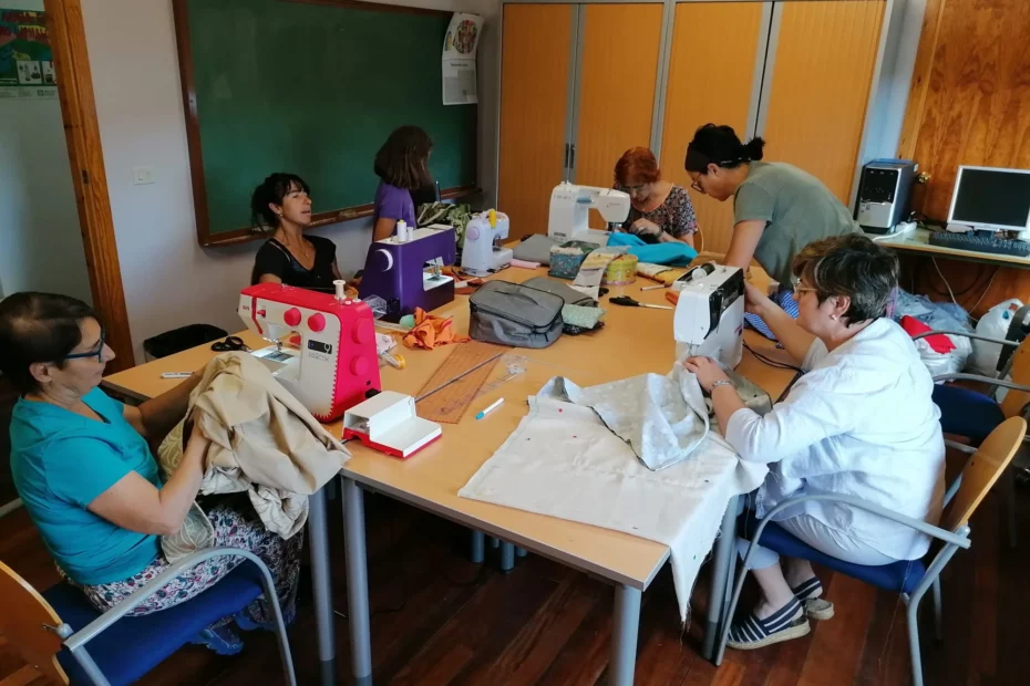 Varias personas realizan el taller de costura. Se ven a seis muejres dentro del aula y alrededor de una mesa en la que hay cinco máquinas de cosas, en las que hay cinco mujeres realizando trabajos de costura con distintas prendas. Además hay otra mujer, junto a la mesa, pero de pie. En la mesa se distinguen distintos objetos como bolígrafos, tijeras, telas, reglas de medir y costureros. En el aúla hay también una pizarra, un armario empotrado y un ordenador.