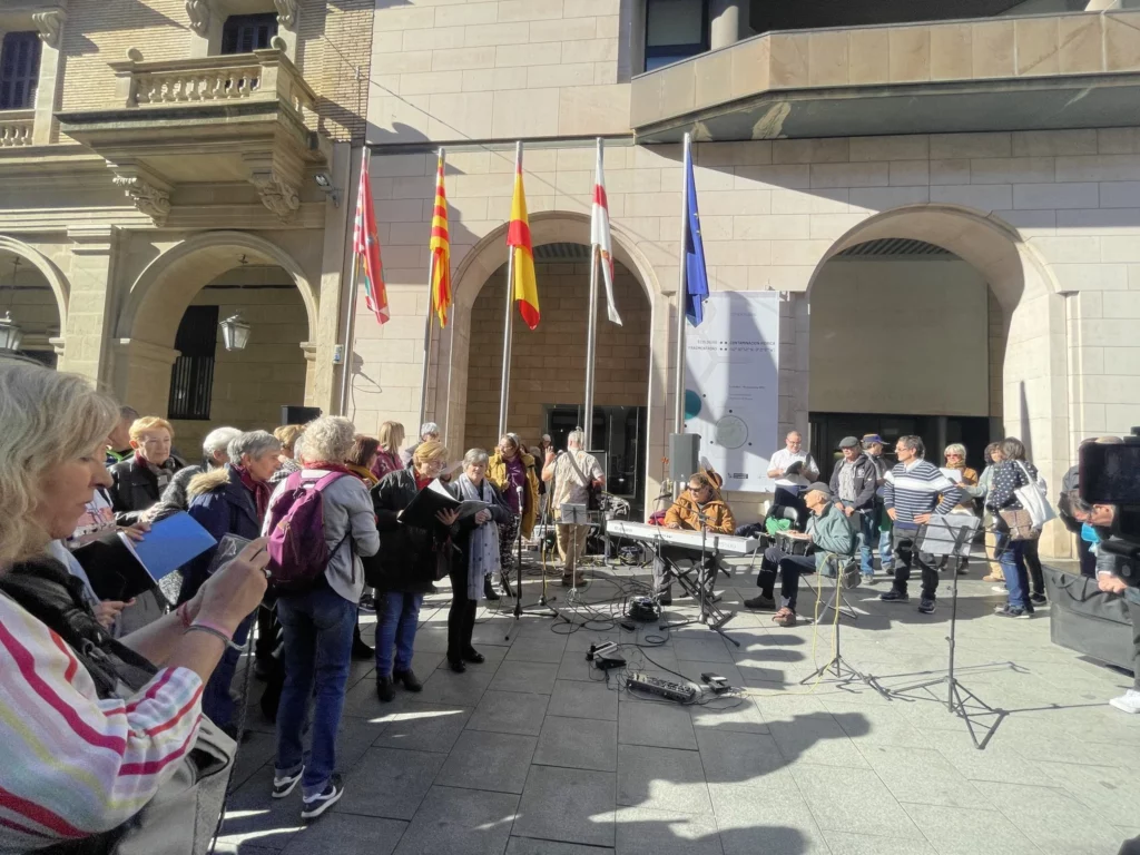 Miembros de la Remós Band actúan en los Porches de Galicia de Huesca, frente a la sede de la Diputación Provincial de Huesca. Junto al grupo hay varias personas pertenecientes al coro Arcadia de la Fundación Agustín Serrate que están revisando las letras de las canciones para cantarlas. Al fondo hay otro grupo del coro y personas del público.