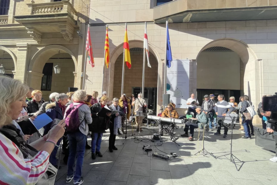 Miembros de la Remós Band actúan en los Porches de Galicia de Huesca, frente a la sede de la Diputación Provincial de Huesca. Junto al grupo hay varias personas pertenecientes al coro Arcadia de la Fundación Agustín Serrate que están revisando las letras de las canciones para cantarlas. Al fondo hay otro grupo del coro y personas del público.