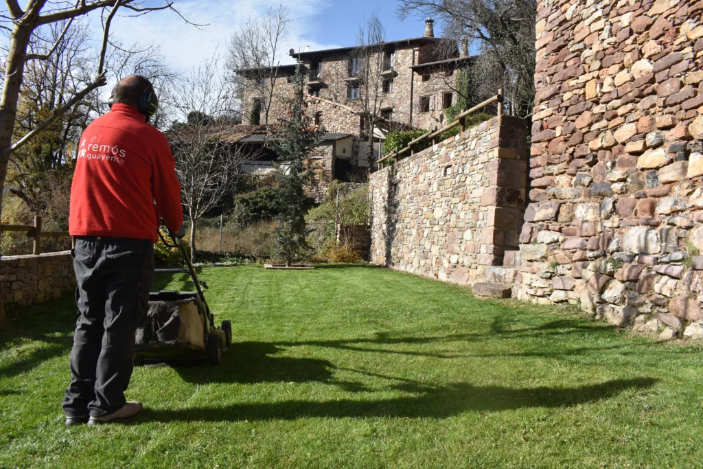 Operario del Centro Especial de Empleo pasa la máquina de cortar césped por un jardín en un día soleado. Al fondo se ven varias casas y a la derecha un muro del edificio.